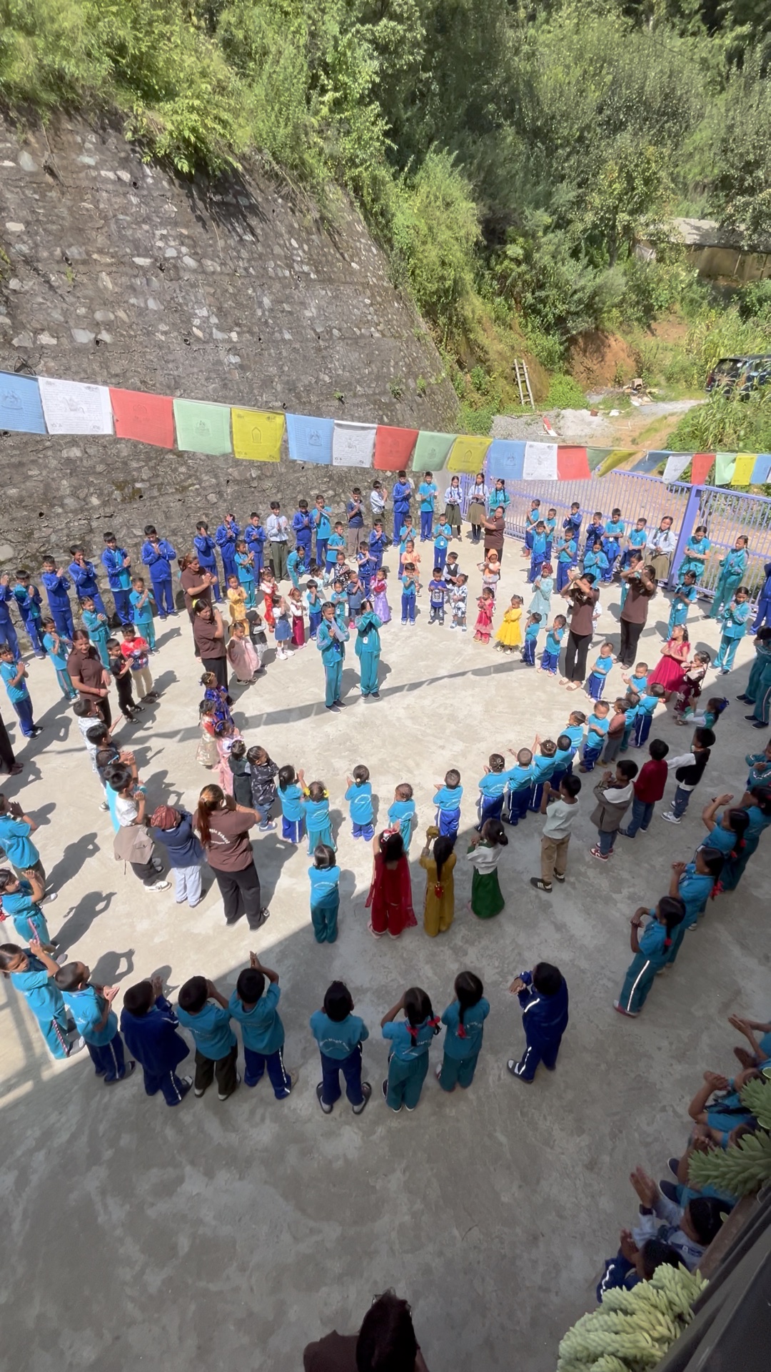 Schultag im Hof der Dolphin Magic School: Kreis der Klasse, Gebetsfahnen, Hangwald hinter der Steinmauer.